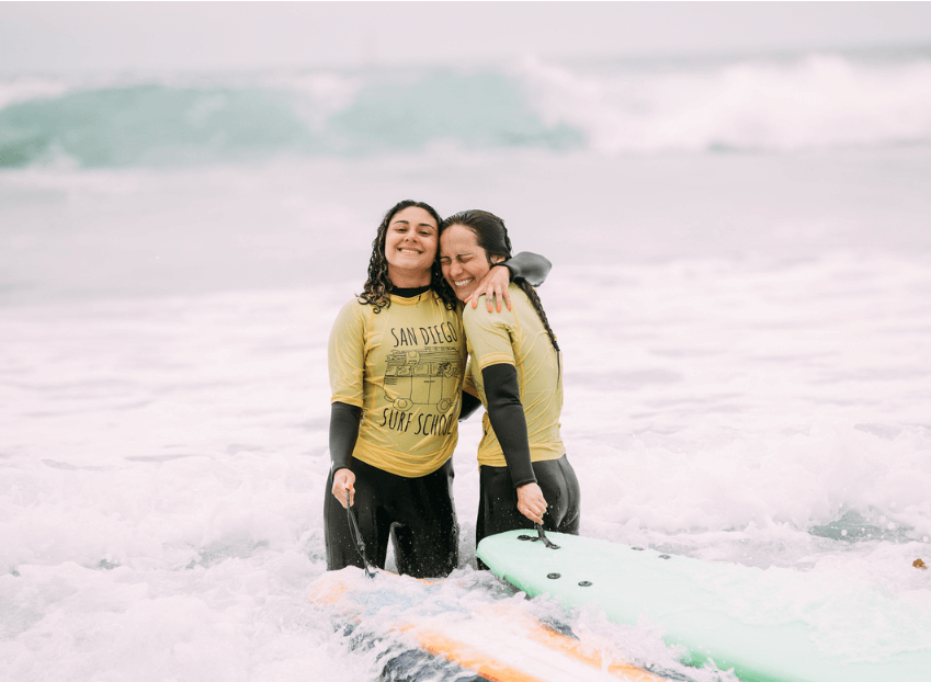 Two women in wetsuits posing beside surfboards on a beach, ready to hit the waves.